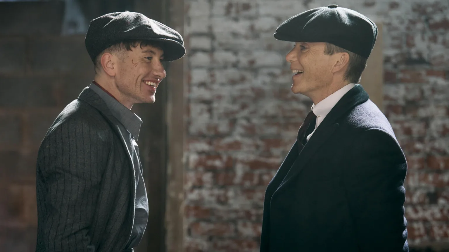 Netflix Barry Keoghan (left) and Cillian Murphy in grey suits and flat caps in a scene from the film. They are both laughing and behind them a brick wall can be seen.