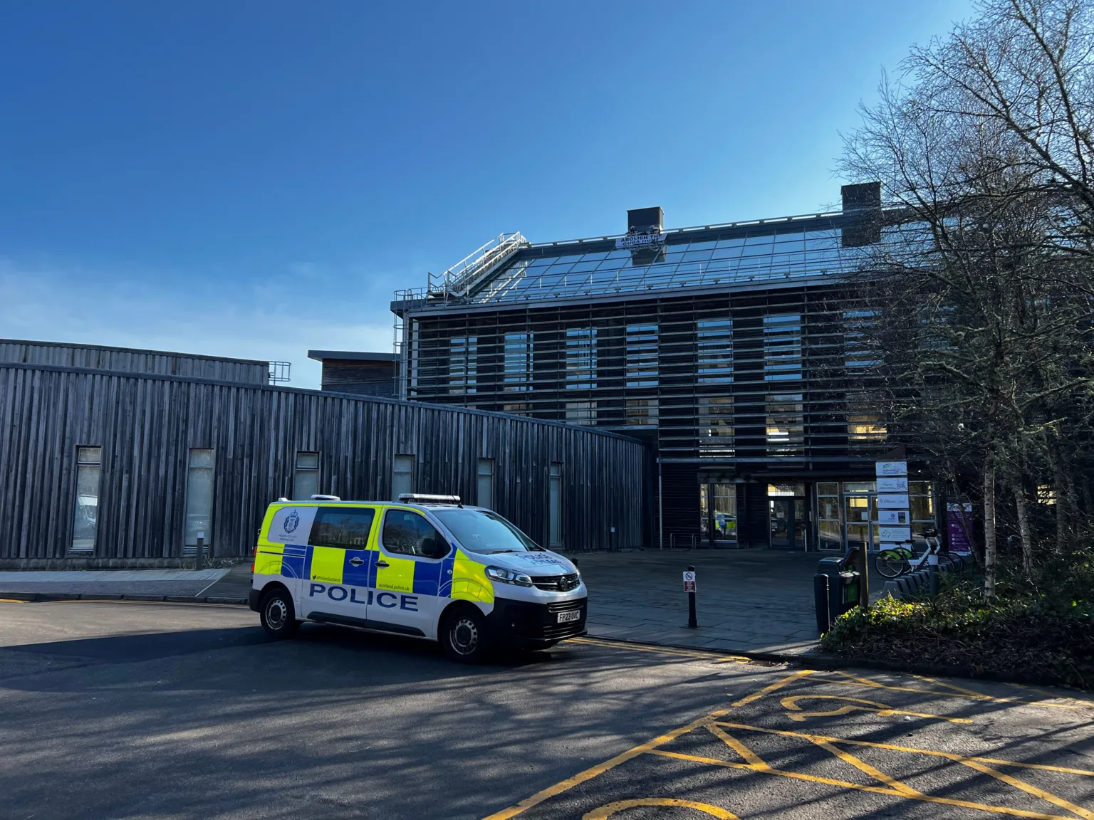 A white police van with yellow and blue markers is parked in front of a larched building. To the right, is a taller building with glass roof in front of a clear blue sky. A tree with no leaves occupies the right hand side.