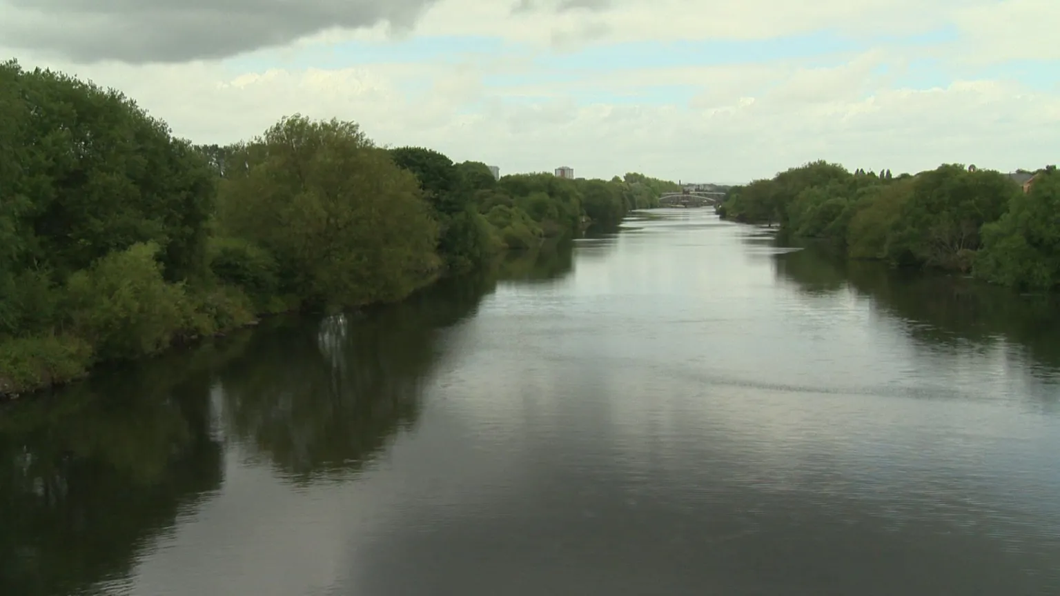 A view of the Manchester Ship Canal with green trees on either side and a view of Salford in the distance