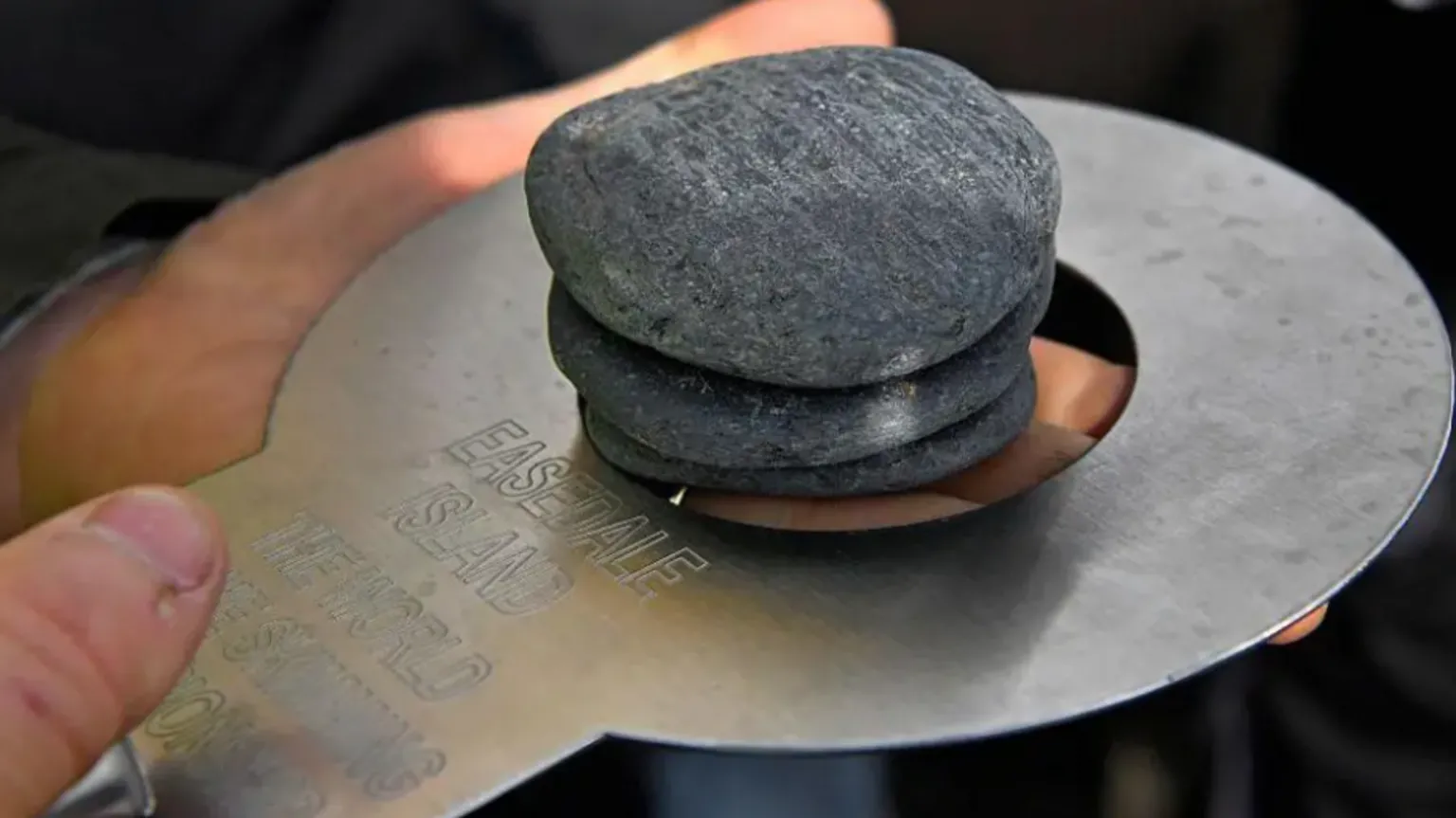 Shows a close-up of a hand holding a metal measuring plate with three flat stones stacked on it, with engraved text visible on the plate.