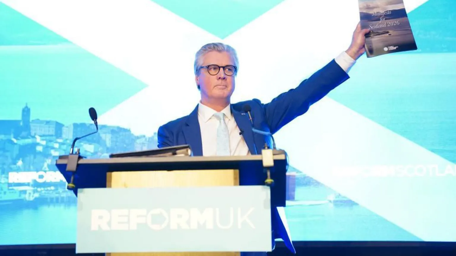 A politician wearing a deep blue suit holding up a manifesto in front of a saltire projected on a screen behind