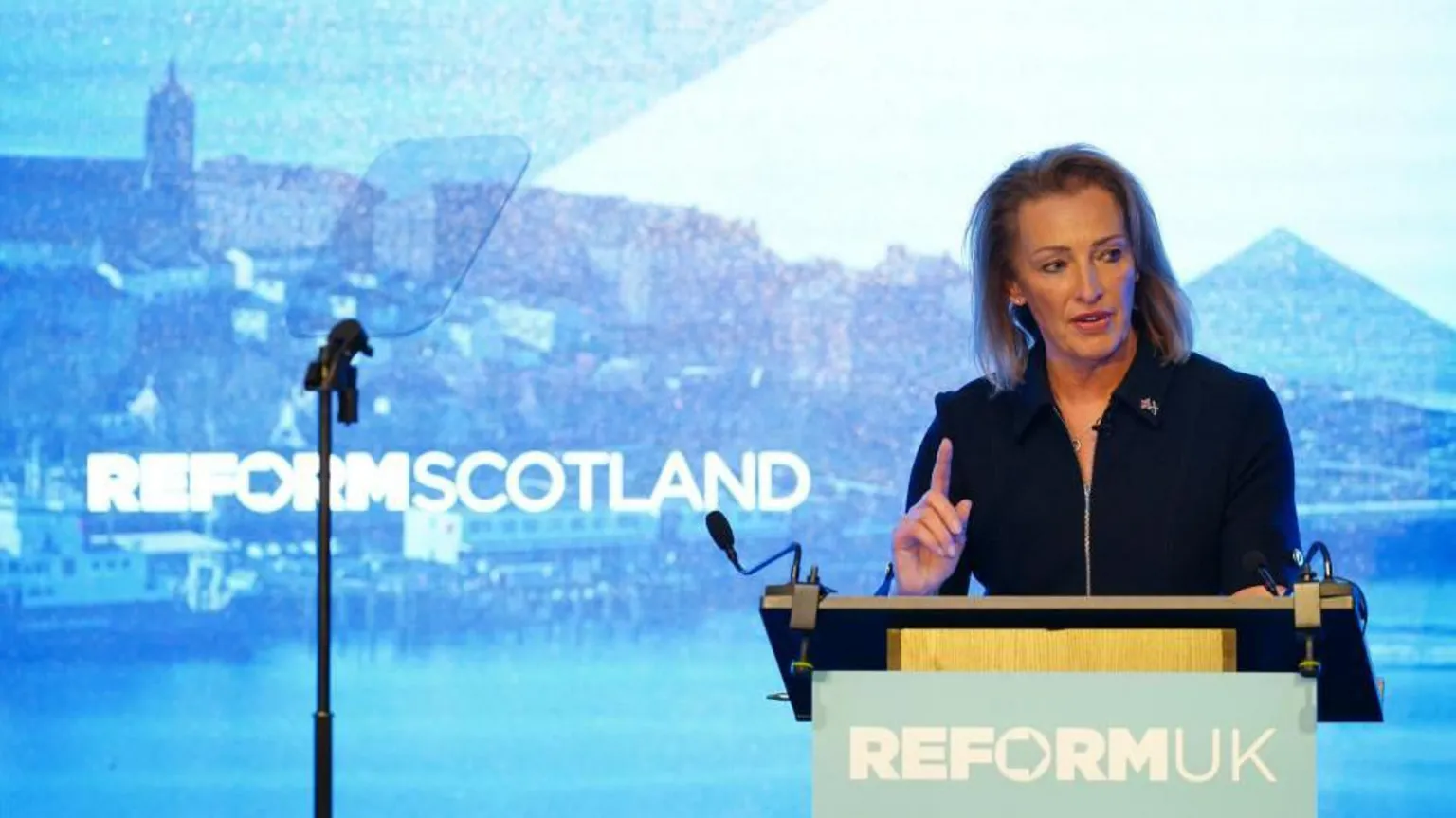 A woman wearing a black suite with her finger pointed in the air stands behind a Reform UK podium