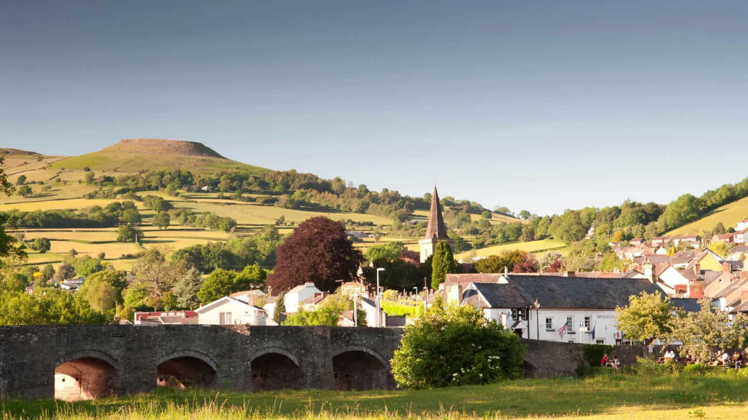 View of Crickhowell seen from across the river with stone bridge with arches in the foreground and table mountain and green hills in the background and the village visible behind the bridge on slightly higher ground, including the spire of a church.