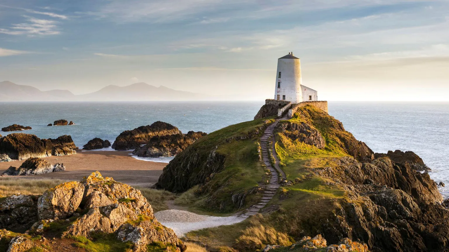 View of Twr Mawr lighthouse on Angelsey surrounded by beach and large rocks on one side and the sea behind, and mountains in the background on the other side of the bay. the lighthouse is a circular white building with steps in the grassy rock on which it is perched leading up to it.