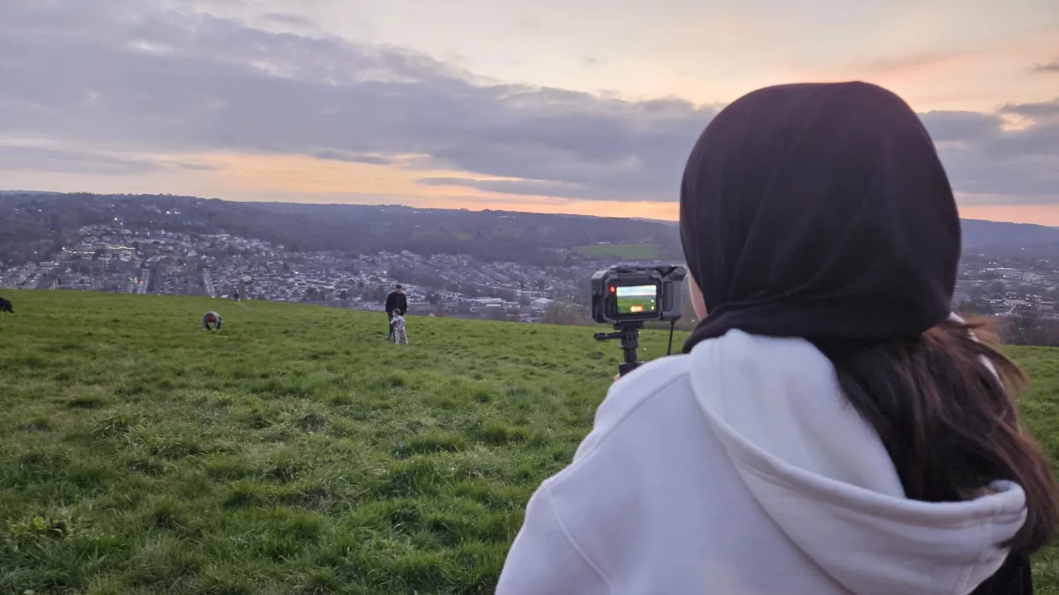 Aisha Khan/BBC A woman with a black headscarf and a white hoodie looks into a camera while standing on a green hill overlooking a city