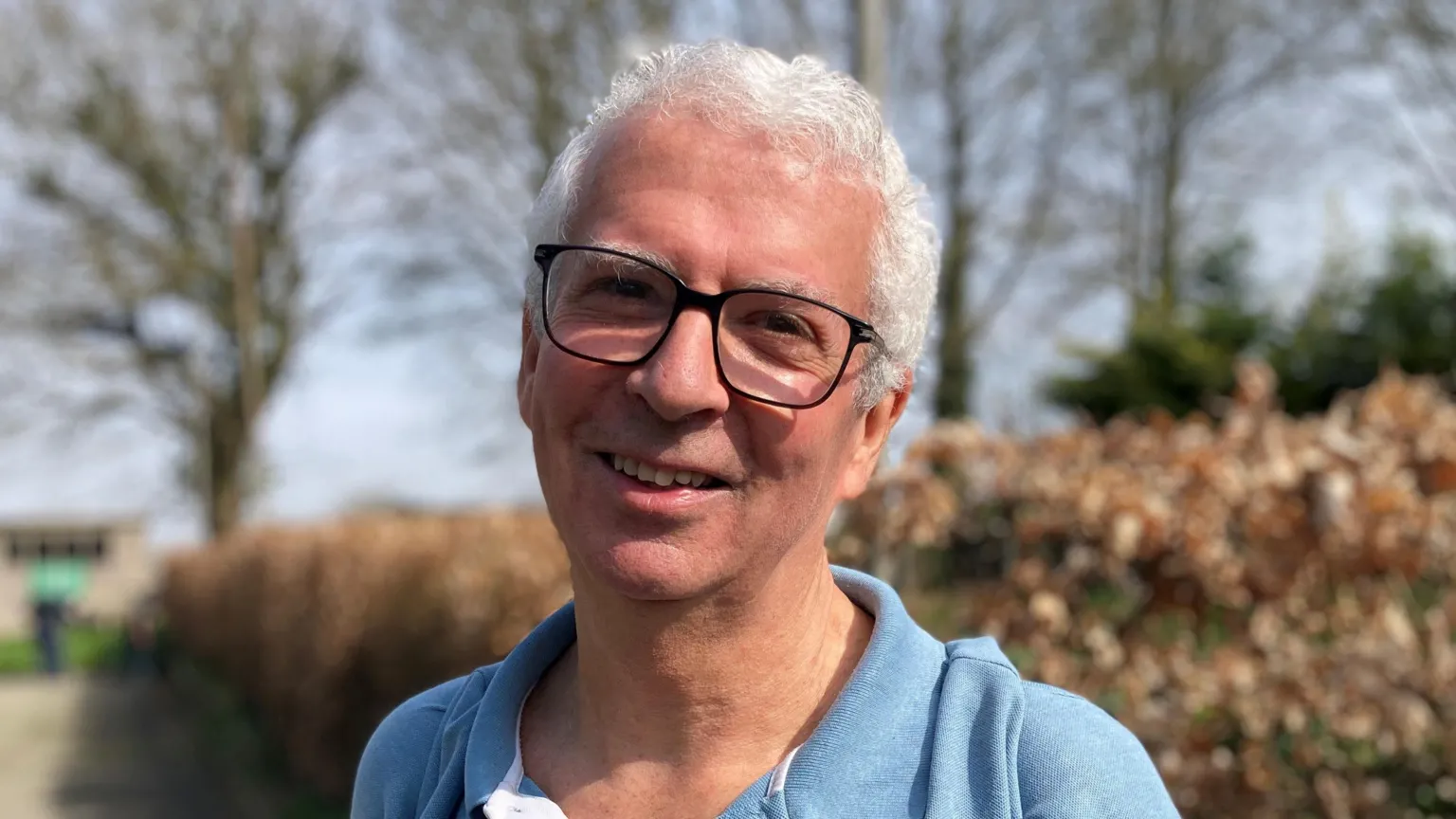 Leo Todd, a white man with short white hair and wide framed glasses, is pictured smiling at the camera. He is wearing a blue collared shirt and is stood in front of a bush. 