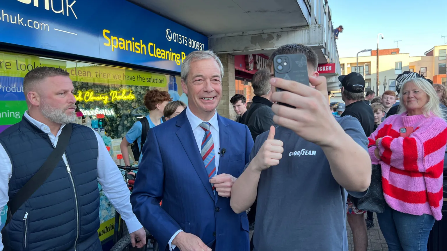 SIMON DEDMAN/BBC Nigel Farage posing for a selfie with a young man holding up an iphone which is covering up his face. Behind him are people who have come out on the street to see the Reform UK leader. They are standing next to a shop with a sign saying spanish cleaning.