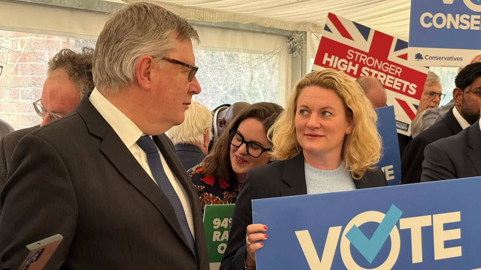 A woman with shoulder length blonde hair holding a big blue placard saying vote looking towards a man to her right with glasses and wearing a dark suit and blue tie. They are at a Conservative party rally.
