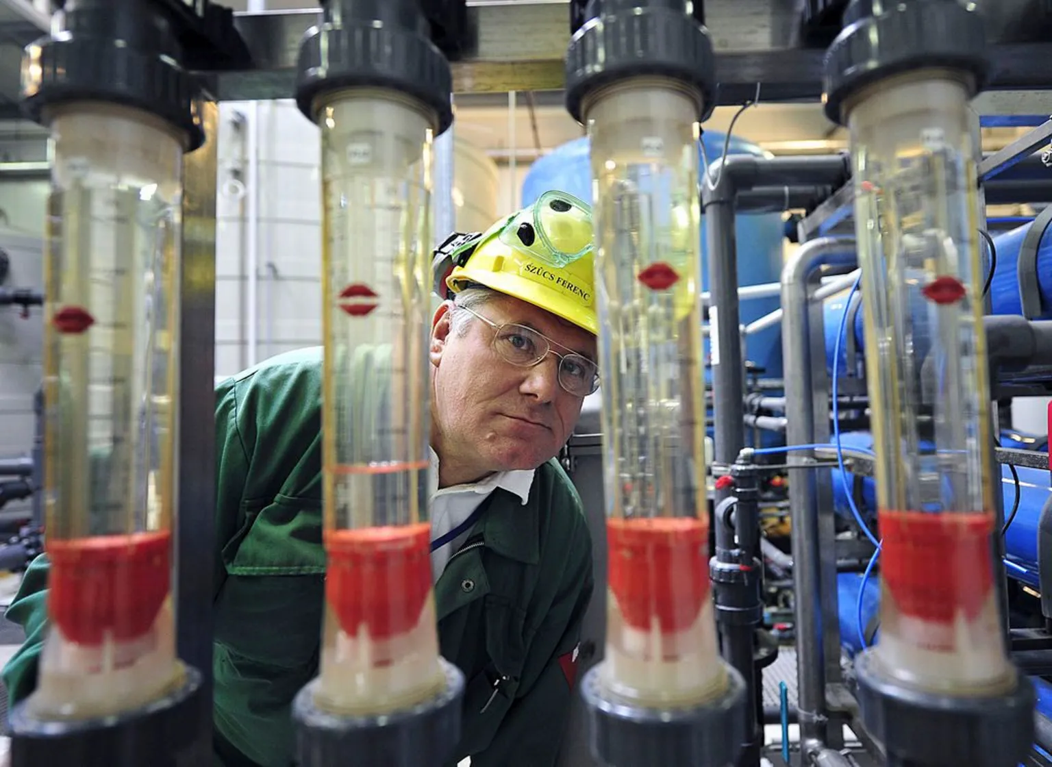 Refinery worker wearing yellow hard hat and green top observes four transparent glass tubes - archive picture