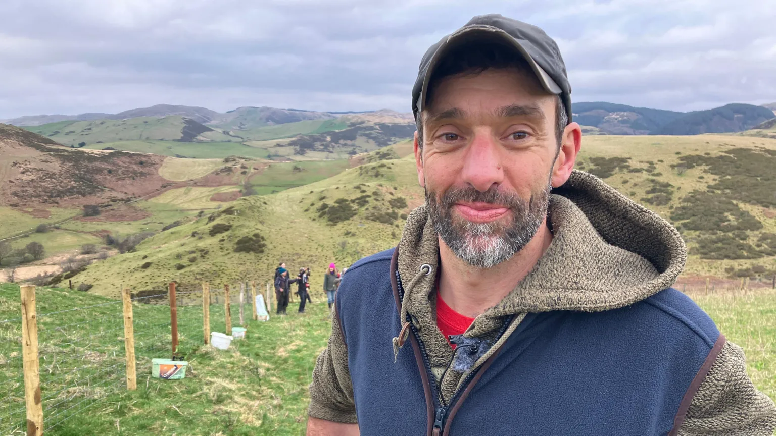A man in his 40's is looking into the camera. He's wearing a cap and has a beard. He's wearing a brown hoodie and a blue gilet. He's standing on a hill. In the backdrop there are a number of hills.