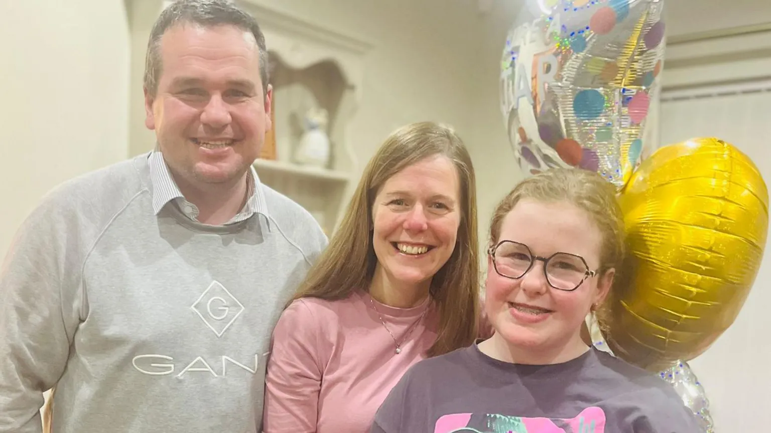 Jennifer Doey Jonny, Jennifer and Lyla are pictured together. Jennifer is wearing a pink top and has long brown hair. Jonny is in a grey sweatshirt and has short black hair. The three are standing near a bunch of balloons.