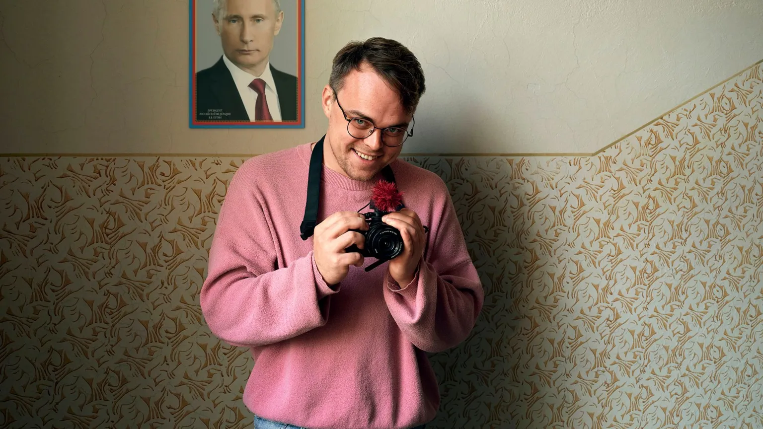 BBC/Made in Copenhagen/František Svatoš A man in a pink sweater holds a camera in front of a picture of Vladimir Putin