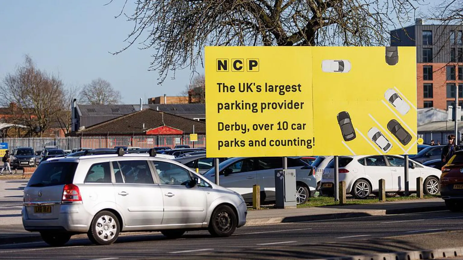 NurPhoto via A silver-white estate car passes by an open-air NCP car park in Derby with a big yellow sign saying