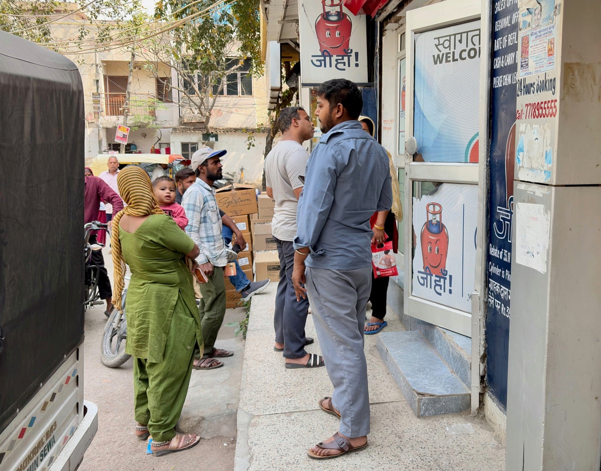 Maya Ranis is seen from behind in the street, holding her baby daughter, as a man stands by. There are signs depicting red gas cylinders and reading Welcome. 