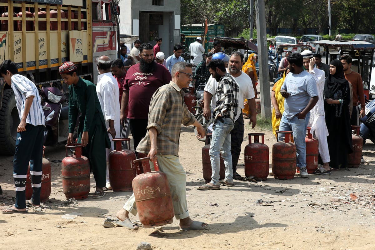 Men and women of varying ages queue in the street with their red gas cylinders; a man in a brown checked shirt carries a refilled one away. 