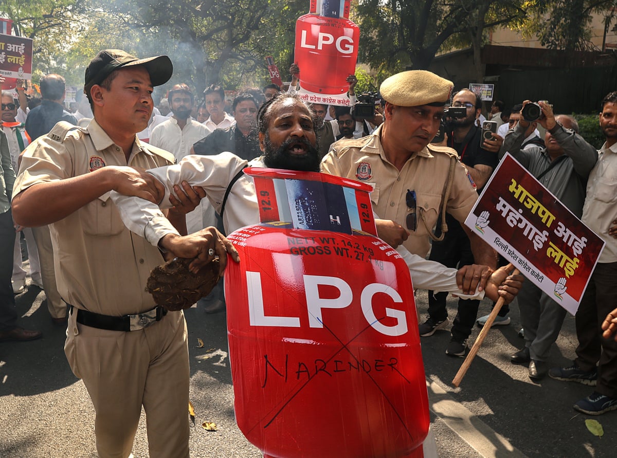 A shouting demonstrator with bushy dark beard is held on either side by police officers. He holds a sign in one hand and a large cardboard cutout of a red LPG cylinder in the other.