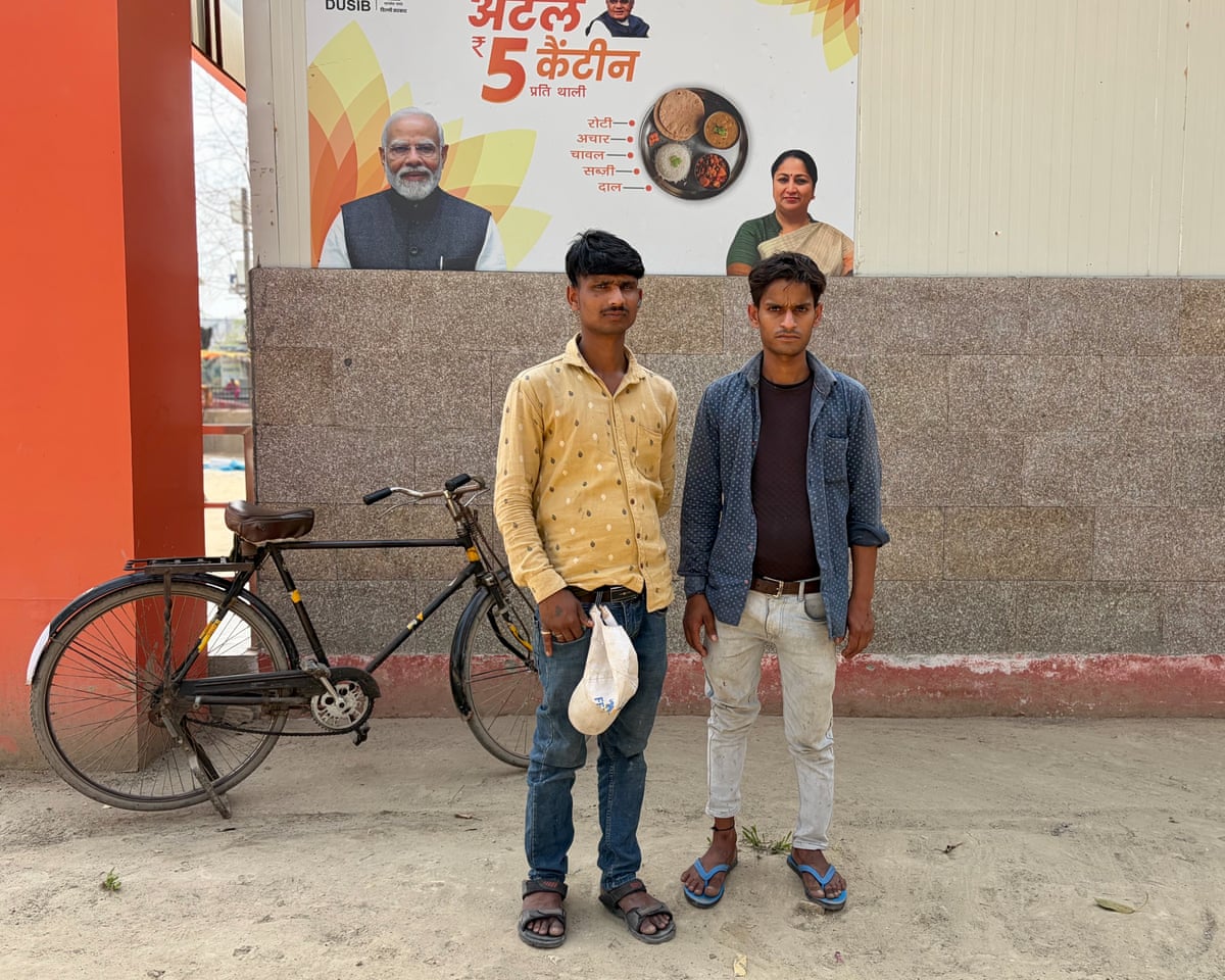Two young men stand by a wall with a poster behind them depicting the Indian prime minister, a bowl of food, and the five-rupee price for the meal. There is an orange pillar and a bicycle behind them. Mandal wears blue jeans and a loose yellow cotton shirt. 