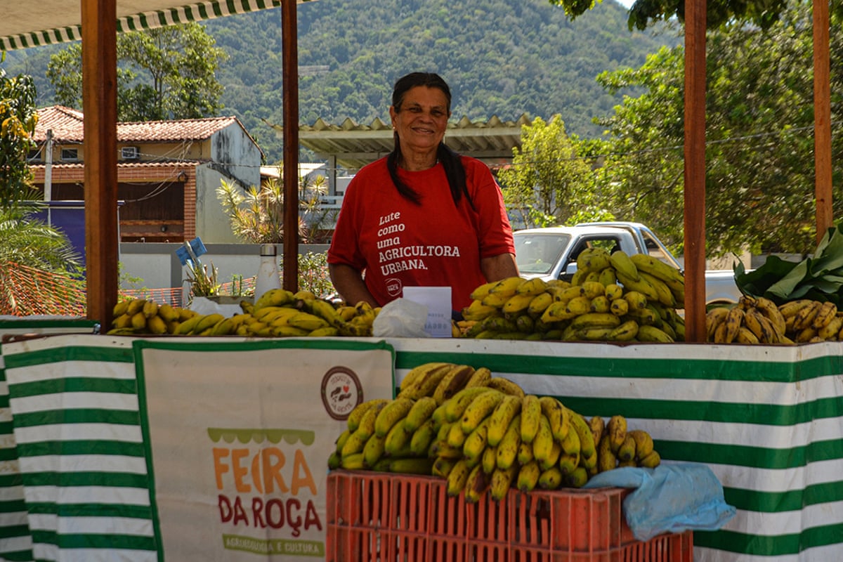 A woman in a red shirt smiles as she stands behind a stall wrapped in green and white striped cloth and piled with bananas.
