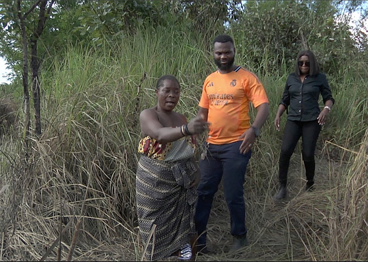 An African woman in a traditional wrap dress, with a man and a woman behind her, points to something off-camera