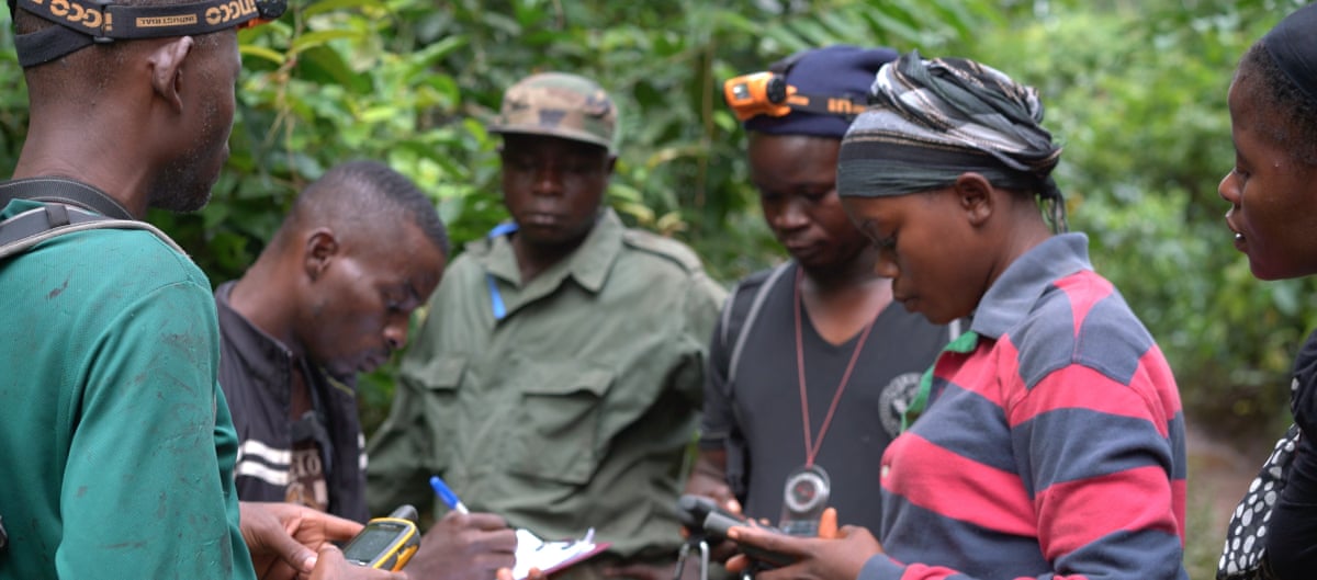 A group of men and women take notes at a briefing in a forest.