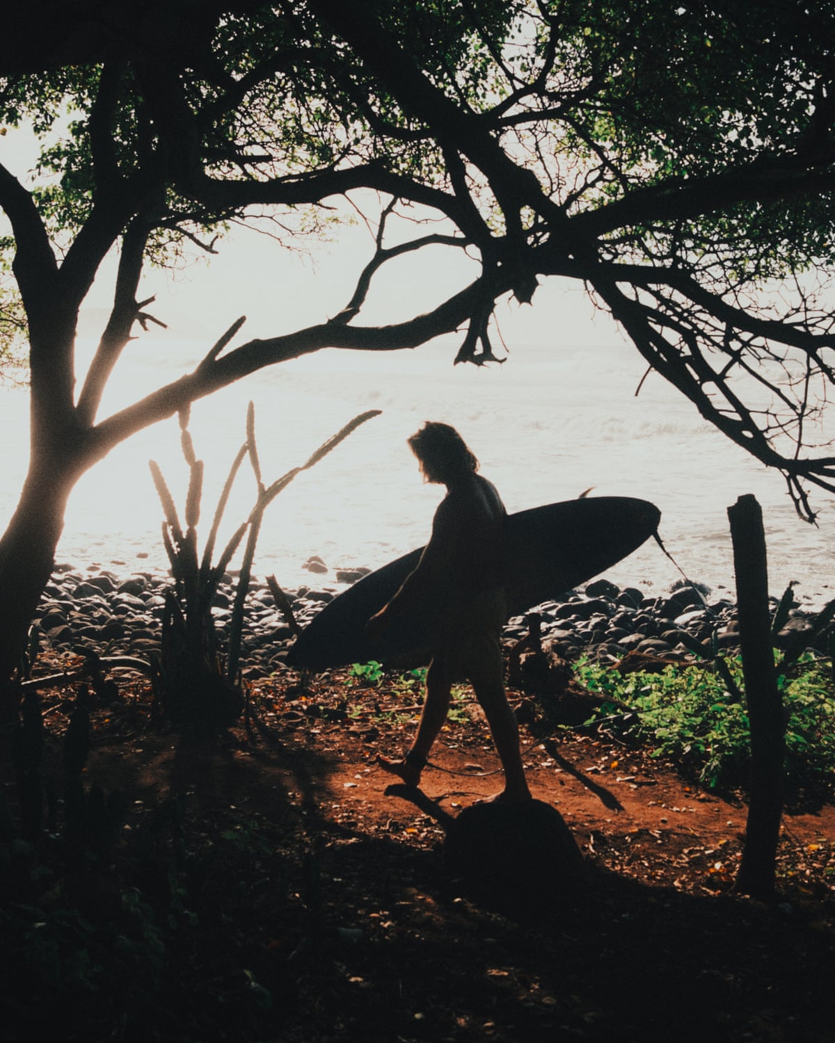 A person carries a surfboard along a dirt track with water in the background