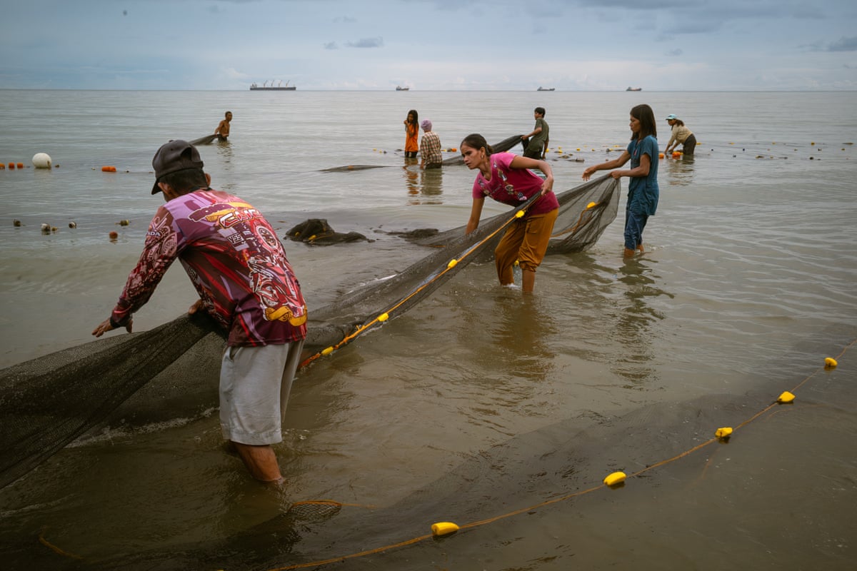 Three people stand in shallow water holding a fishing net. Other people holding nets can be seen behind them