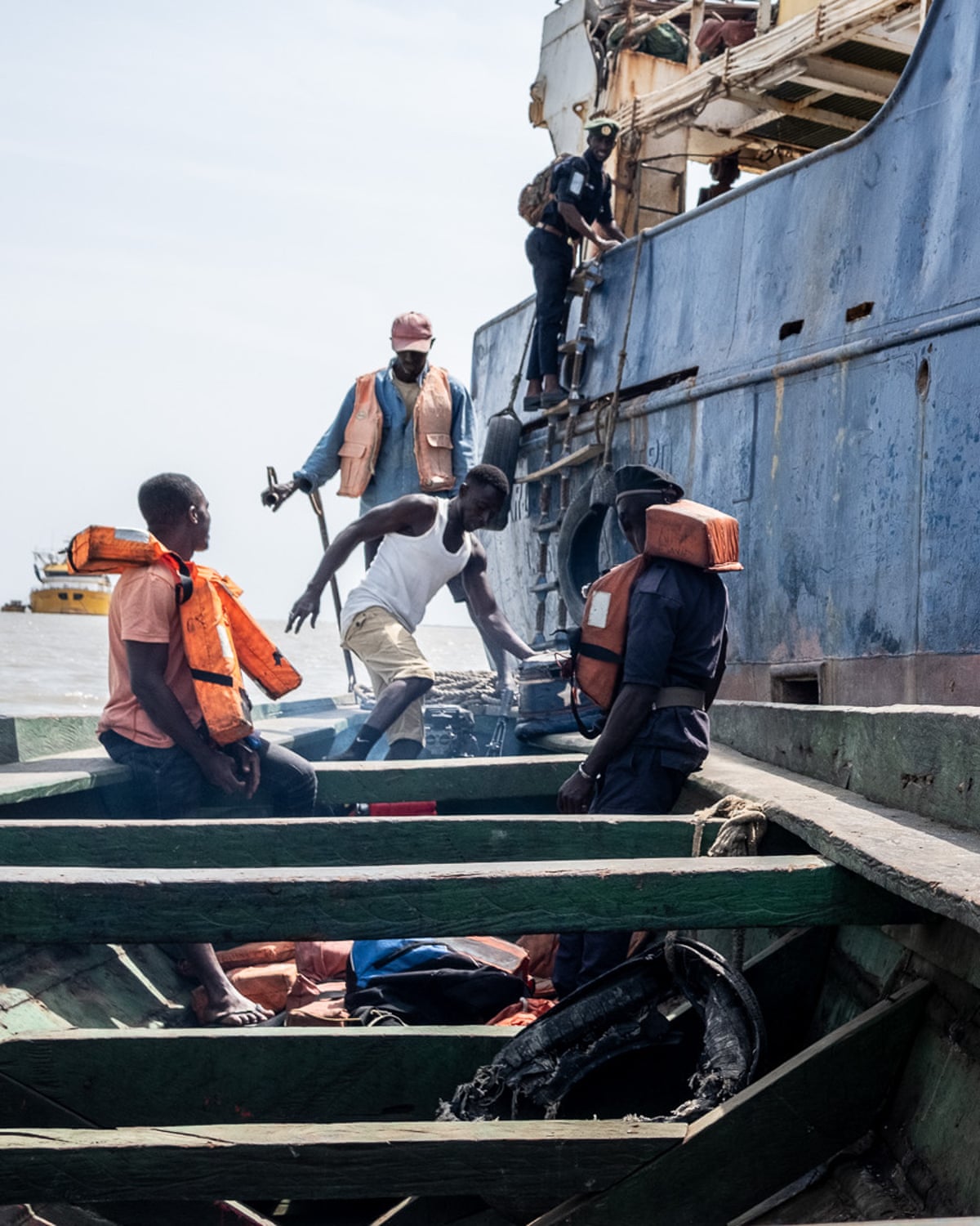 A man in uniform climbs a ladder on the side of a large boat from a smaller boat with four other people in it, one of whom is also in uniform