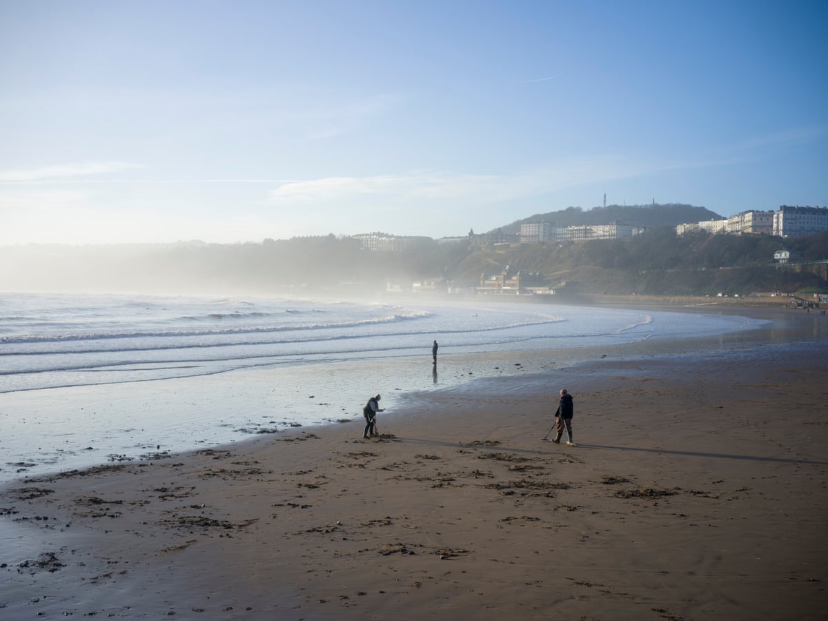 A view of two metal detectorists on a sandy beach in a wide bay with another person in the distance staring out to sea