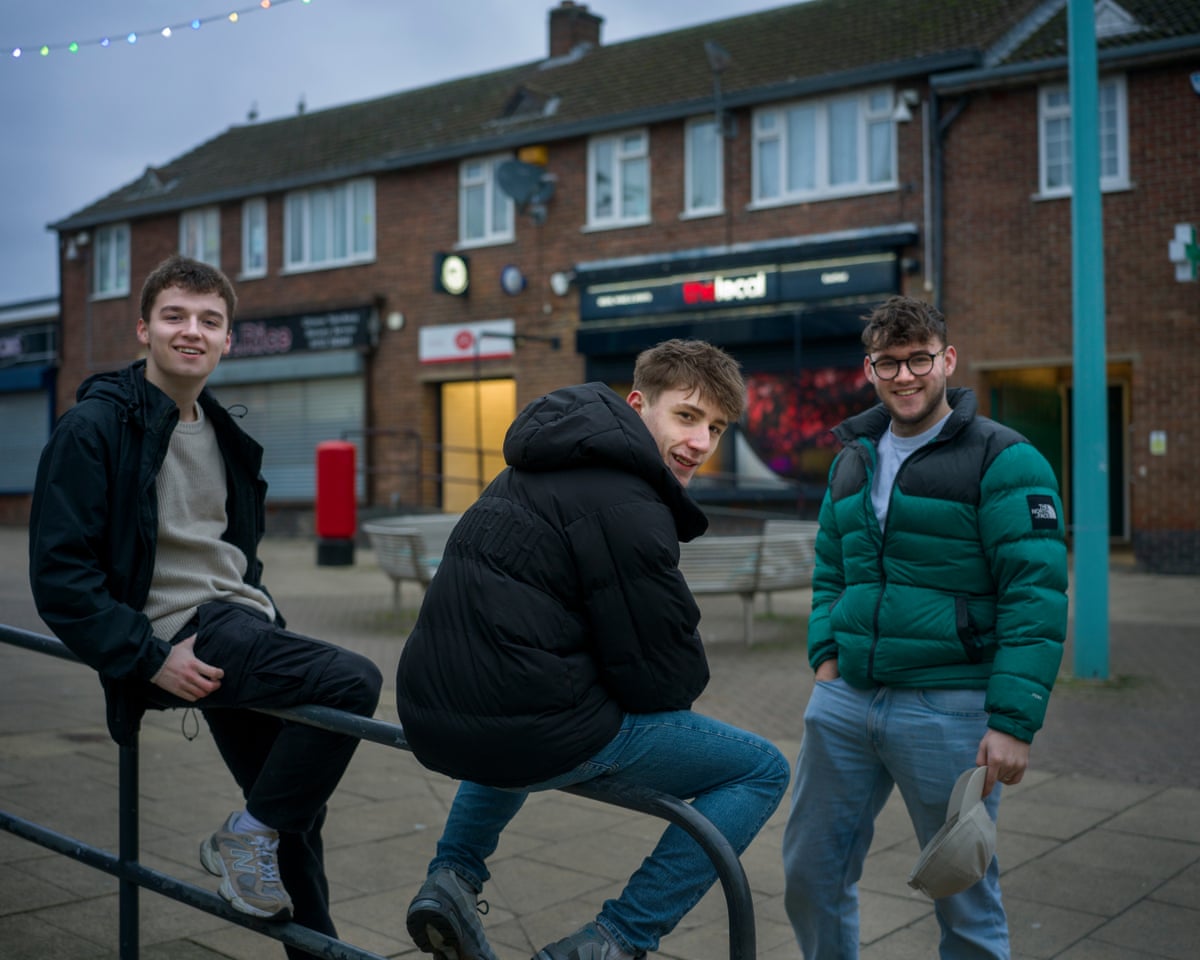 Three teenage lads hanging out on a street by some shops, two sitting on a railing