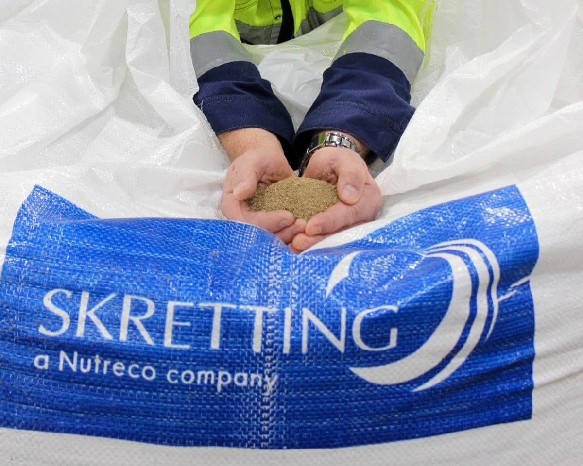 Hands hold a brown pile of feed above what appears to be a bag reading: ‘Skretting, a Nutreco company’