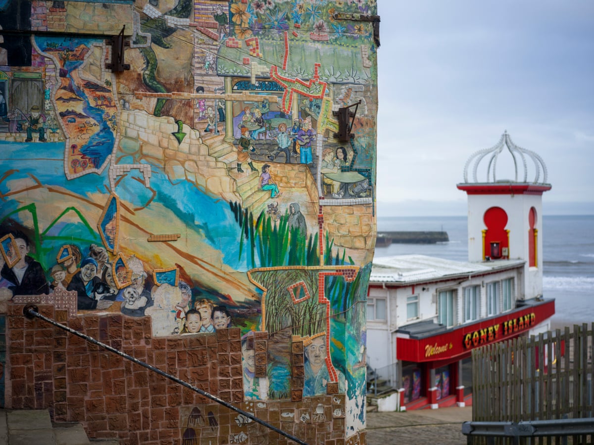 A mural featuring children’s and other faces, and scenes from the town on a wall alongside steps leading to an old-fashioned arcade on the seafront
