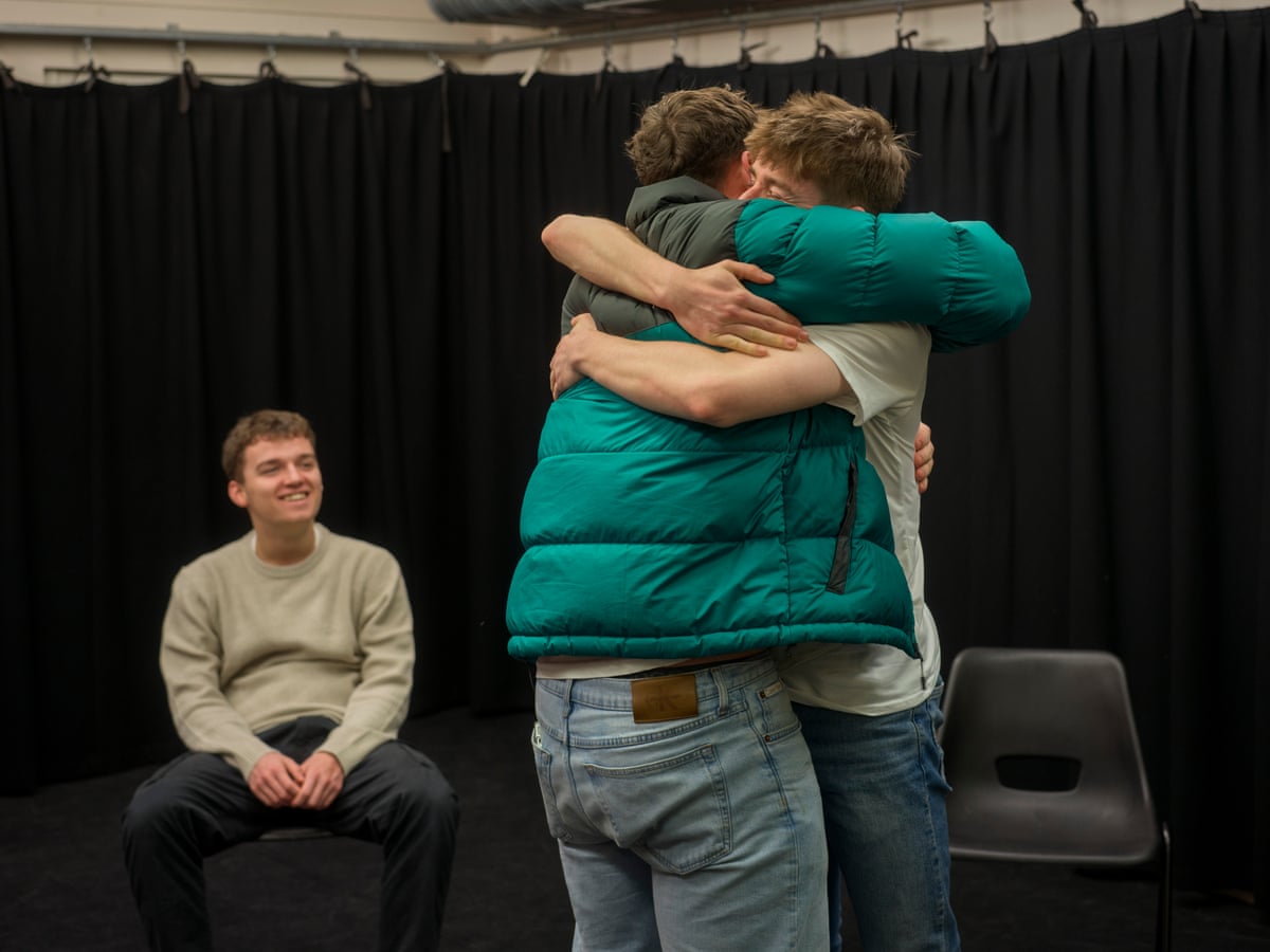 A seated teenage boy smiles as he watches tow other teenagers hug each other in a rehearsal space 
