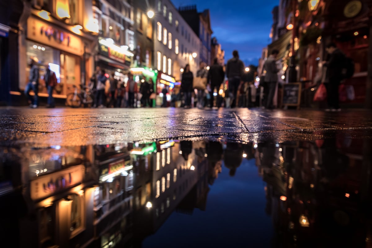 A night image of a streets in London’s Soho district on a rainy evening with the shop lights reflected in a puddle.