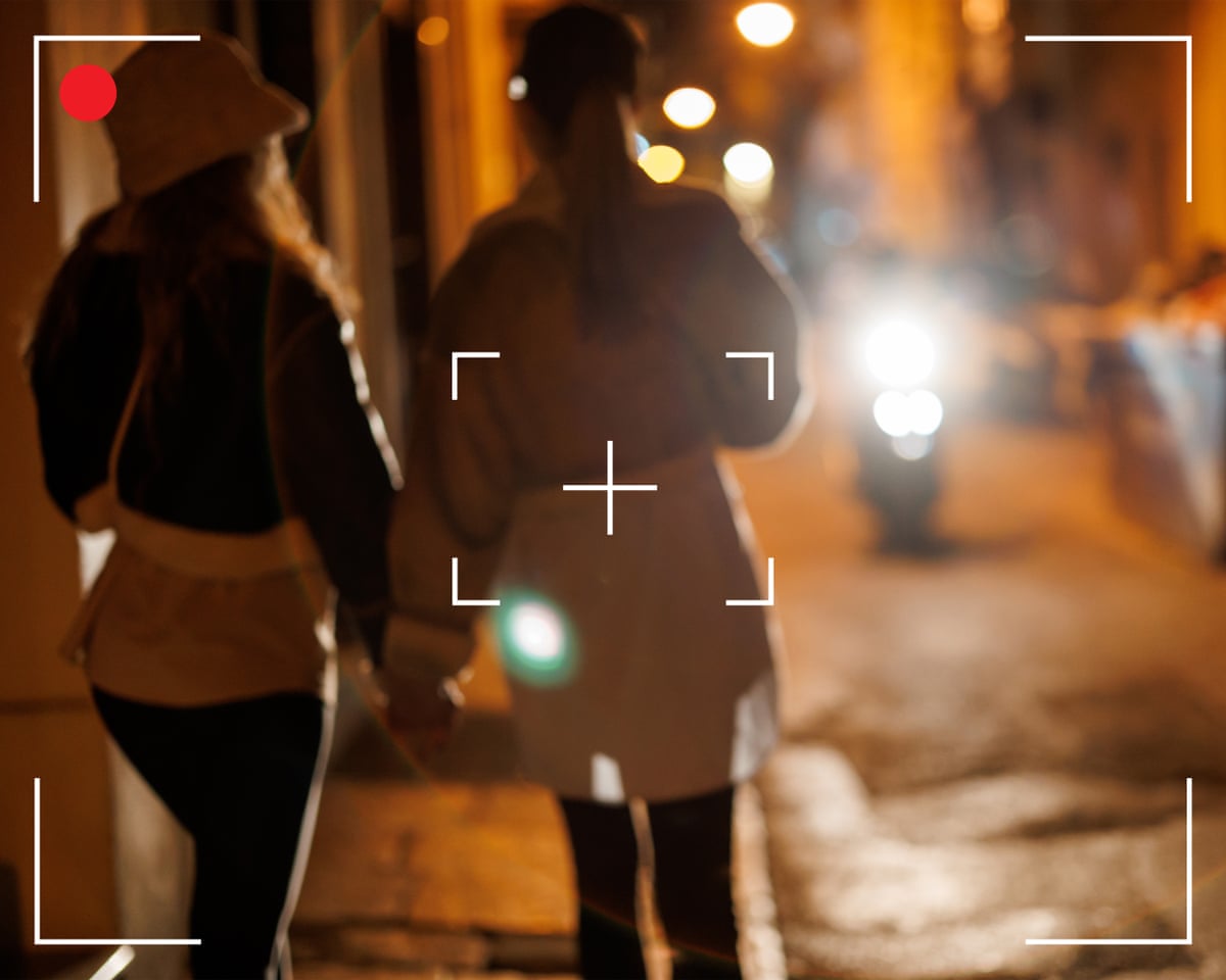 Back view of two women holding hands and walking in city at night with a camera viewfinder frame superimposed