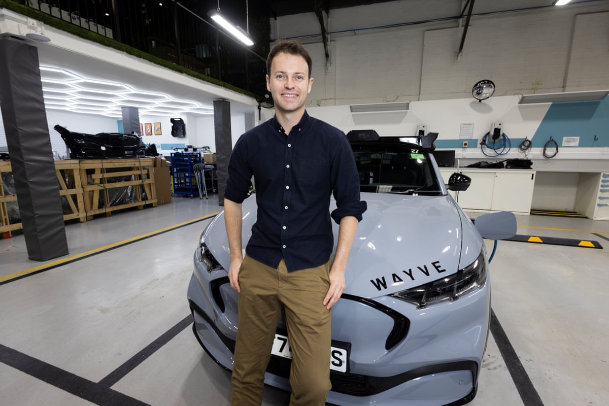 Wearing a navy button-up shirt and khaki trousers, he poses, smiling, in front of a grey Wayve automated vehicle in a factory setting