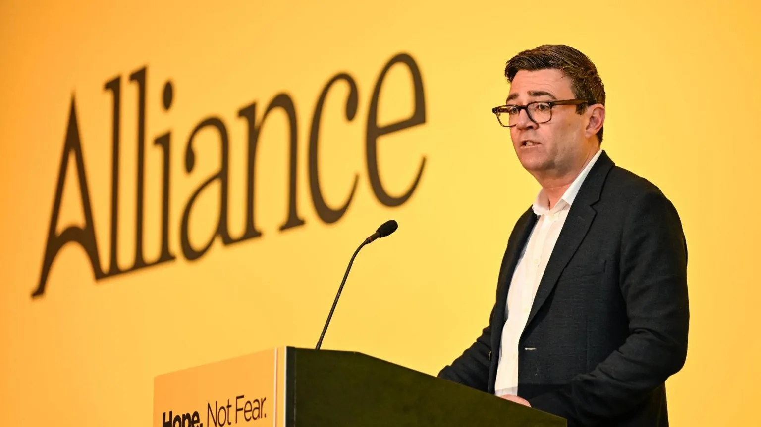  Andy Burnham stands up on stage in front of a large yellow Alliance banner. He is stood at a podium with a singular long microphone. He has short, brown hair and is wearing a dark jacket and a white shirt.