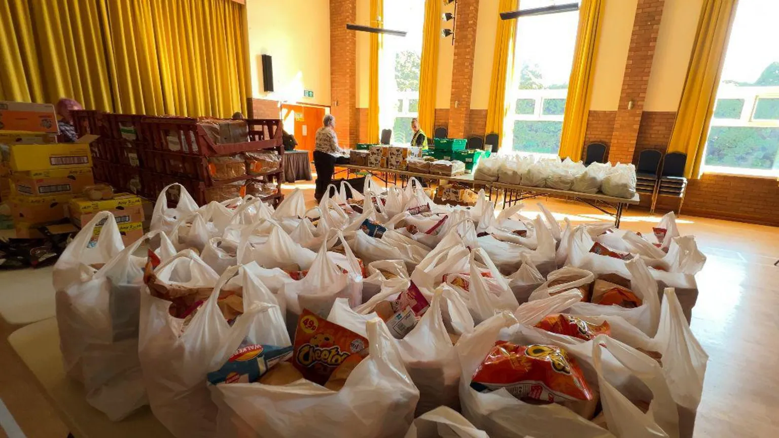 A community hall with two tables. On each table is dozens of white plastic bags filled with food, from packets of crisps to fruit. Next to the bags are pallets of bread, cakes and biscuits. People can be seen in the background organising the food. Light floods in through tall windows, highlighting a wooden floor and tall, yellow sets of curtains.