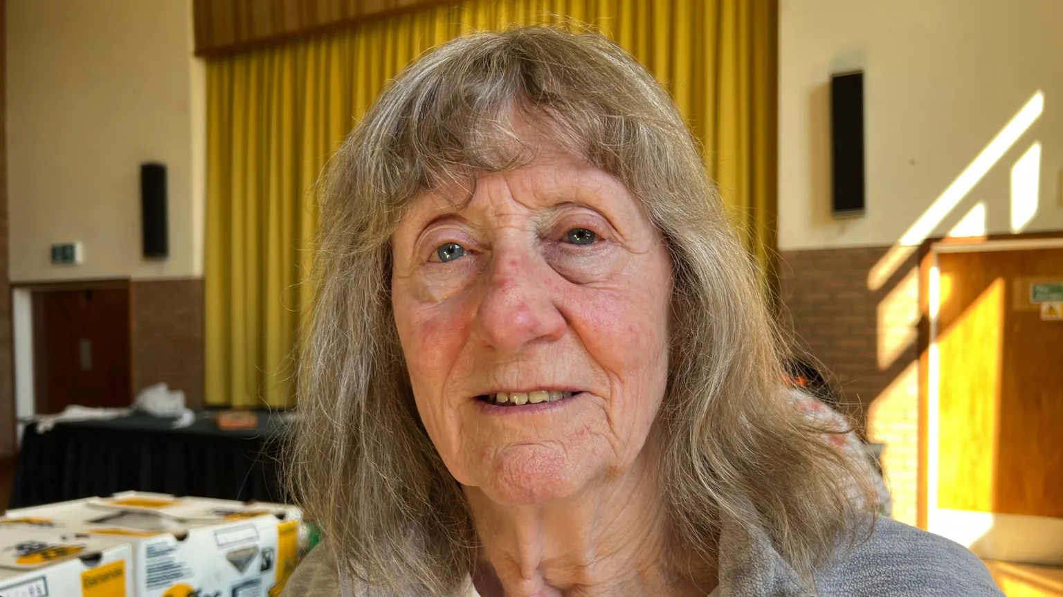 Cheryl Wray has dark-blonde, medium-length hair. She has blue eyes and is smiling. Behind her is the community centre's stage, with an olive green curtain. 
