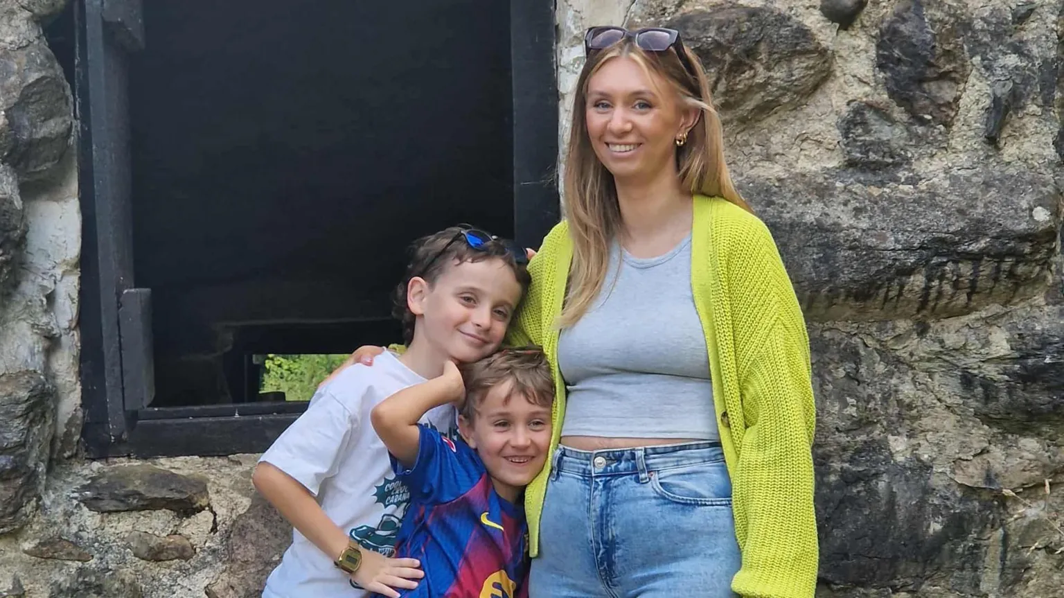 Zoe Stokes-Jones Photograph of Zoe Stokes-Jones (right) pictured with both of her sons. Jacob is on the left, with her youngest son in the middle. She wears a bright green cardigan, a grey t-shirt and blue jeans. She has blonde long hair and sunglasses on her head. They stand outside a stone wall building.