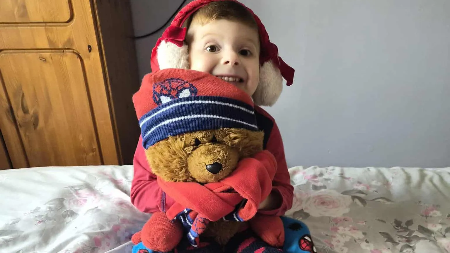 Stacey Angel Brayen, aged five, pictured sat on a double bed in a bedroom. He wears red and white fluffy earmuffs and a red long sleeve t-shirt and smiles at the camera. He holds a large brown stuffed teddy bear, wearing a red and blue spider man hat and scarf. Behind him on the left is a wooden wardrobe, while a grey painted wall covers the rest of the background.