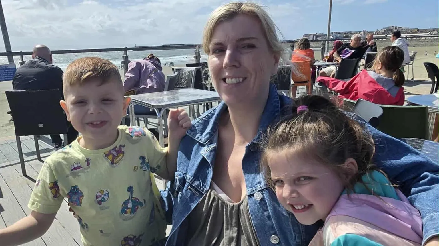 Stacey Angel Photograph of Stacey Angel (Centre) and her son, Brayen, (left) and her granddaughter. Stacey has blonde hair and smiles at the camera. They all sit on a patio looking out over the seaside.