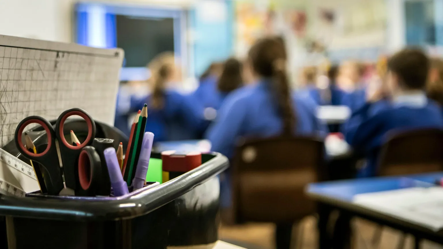 A generic image of a classroom with children in the midground in blue jumpers. There is a board on the wall showing teaching aids and there is a box of pens and pencils in the foreground.