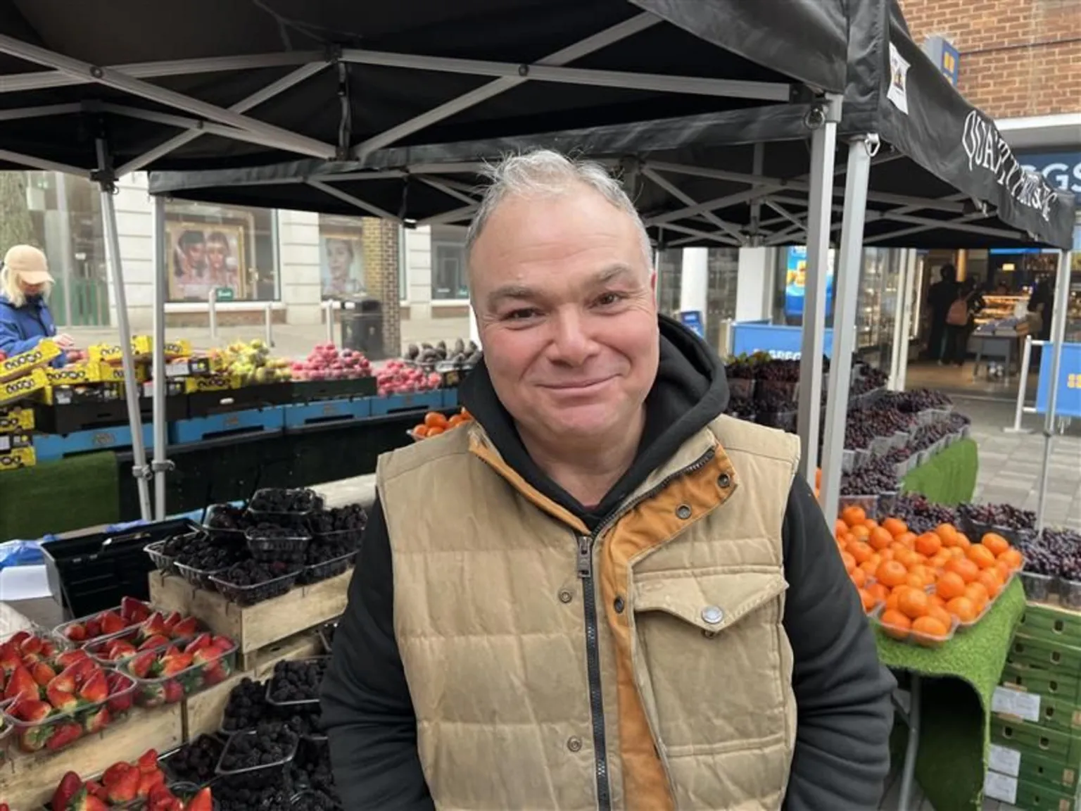 Phil Harrison/BBC A man in a beige jacket smiling for the camera. Several varieties of fruit are stacked up in the background at a colourful fruit staff.