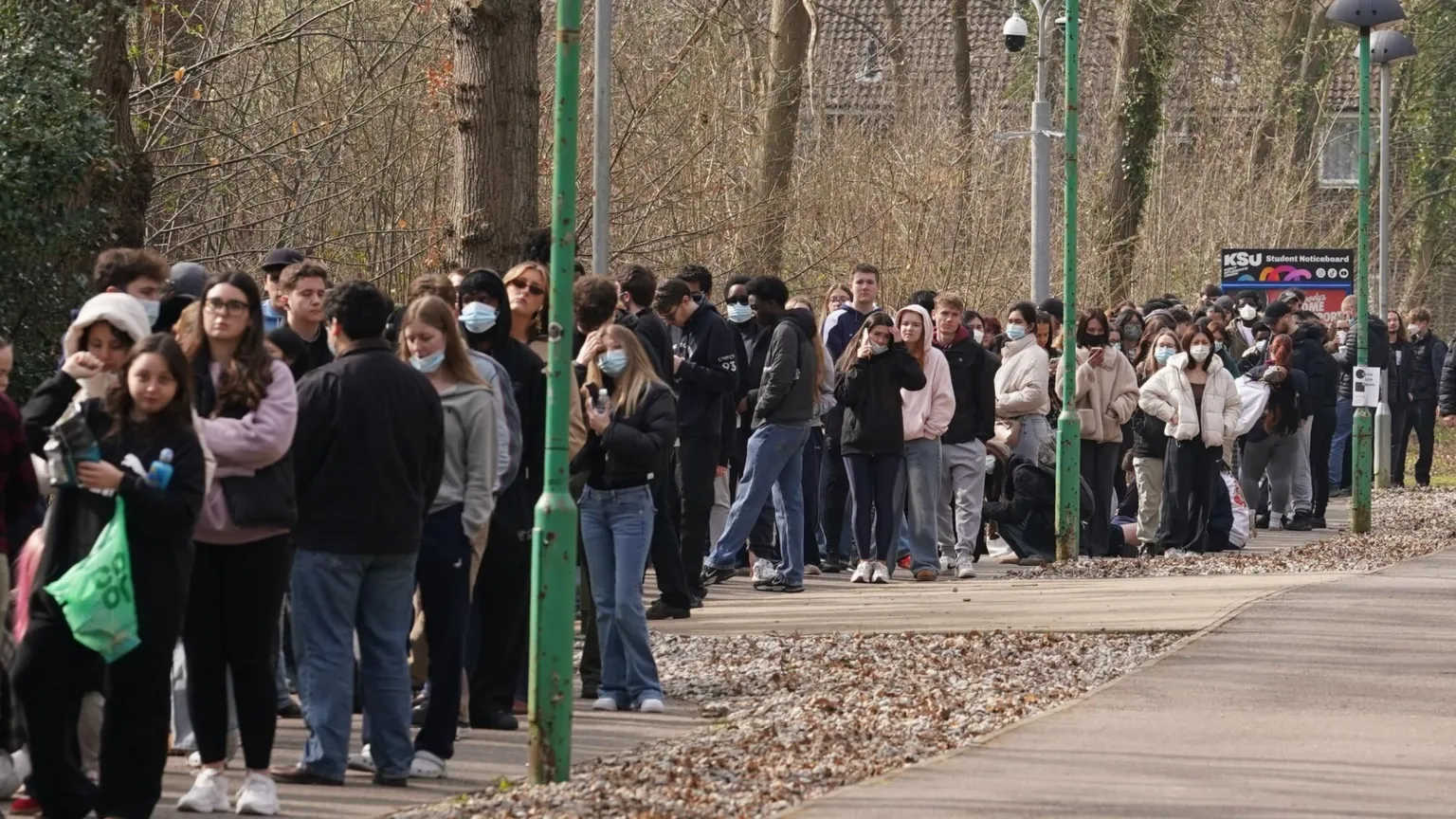  Dozens of young people queue on a concrete path lined with trees. Many are seen wearing face masks.