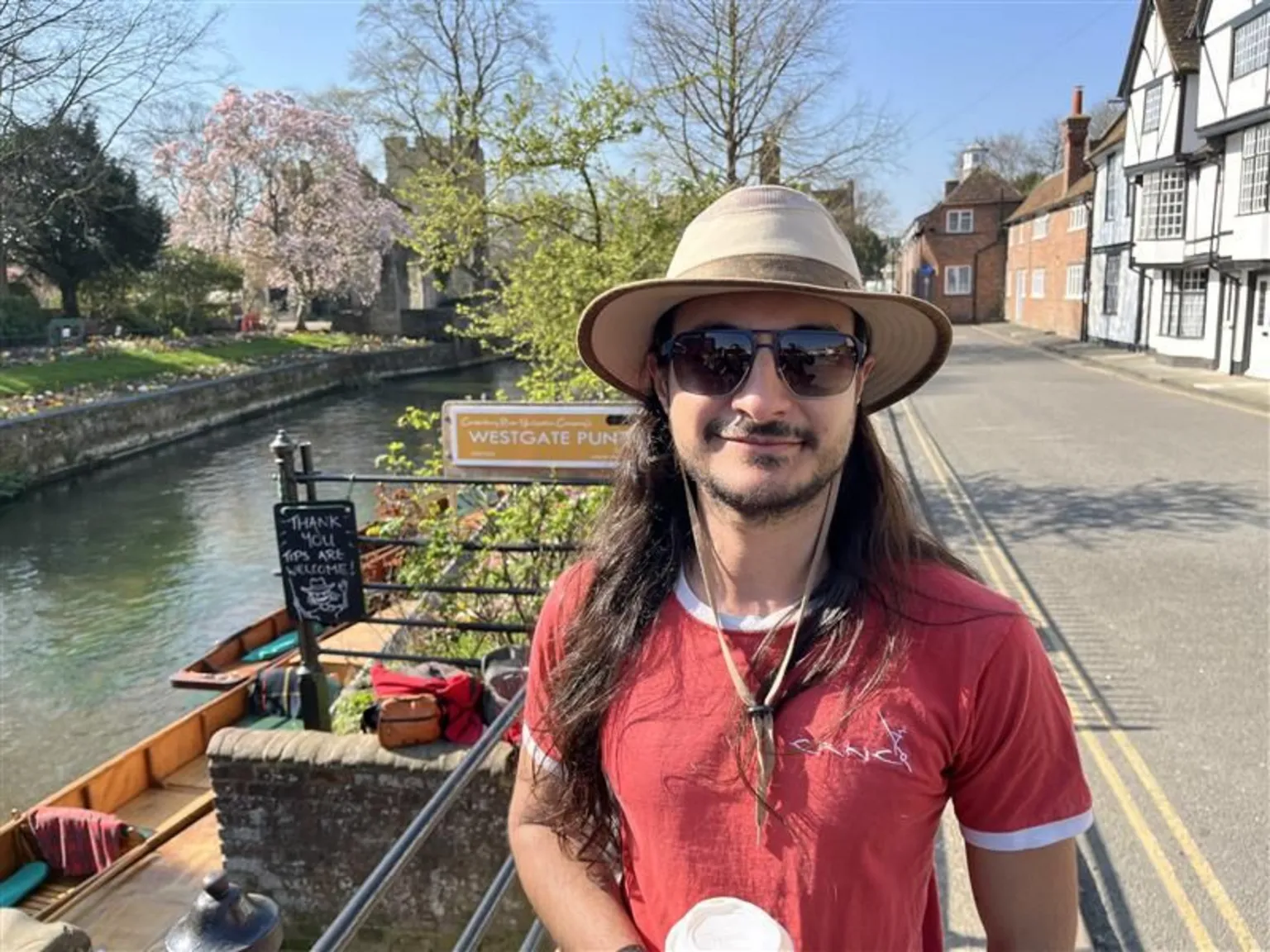 Phil Harrison/BBC A man in a red t-shirt, sunglasses and a wide-brimmed hat smiling at the camera. A river with some wooden punt boats at the side of the water is seen behind him. It is a sunny day.