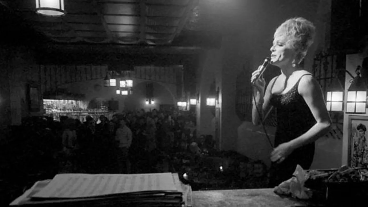 Watford/Mirrorpix/Getty A black and white photo of drag queen Shane performing downstairs at The Black Cap. She is stood on the stage on the right, wearing a strapless dress, hair up in a chignon, and in the background is the audience. She is holding a wired mike and the spotlight is shining on her.