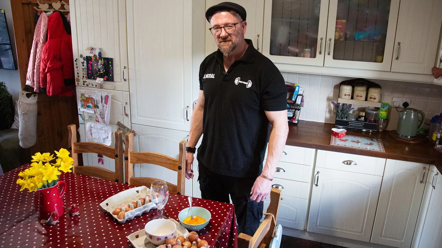 A person stands in a brightly lit kitchen beside a table, where there are several bowls and egg cartons. The person is wearing a dark polo shirt with a small weightlifting graphic and text on the chest.