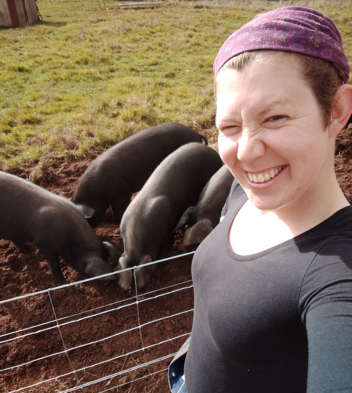 A woman, Lucy Ridge, in a purple headband and a black top, taking a selfie on a farm. In the background are three black pigs