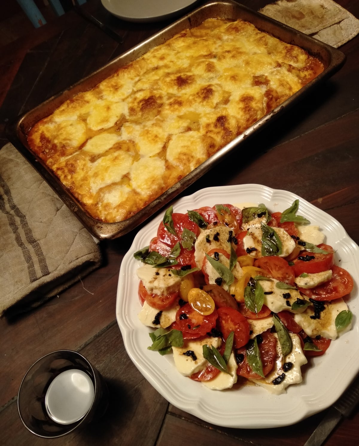 A colourful tomato salad on a white plate and a tray of lasagne on a wooden table