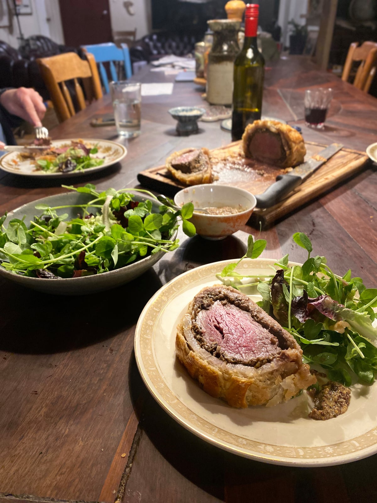 A long wooden table, set for a meal. In the foreground is a white plate with a slice of beef wellington with a leafy salad. In the background is a bowl of salad, and a cutting board with a knife and more beef wellington